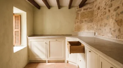 Cream painted cabinetry scribed to an uneven plaster wall under a stone worktop, one oak drawer open, a terracotta floor