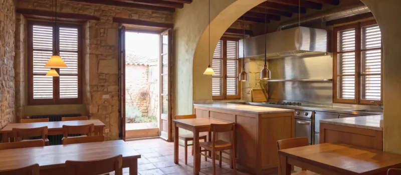 Comedor de restaurante con mesas de madera y un arco de piedra hacia una cocina de acero, persianas cerradas y lámparas colgantes encendidas