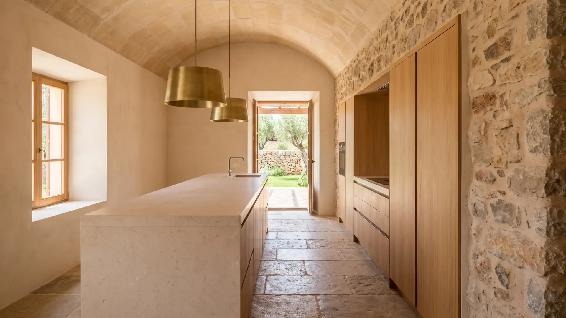 Gewölbte Finca-Küche mit einer langen Kalkstein-Kücheninsel und bündigen Eichen-Hochschränken in einer Bruchsteinwand, zwei Messingleuchten
