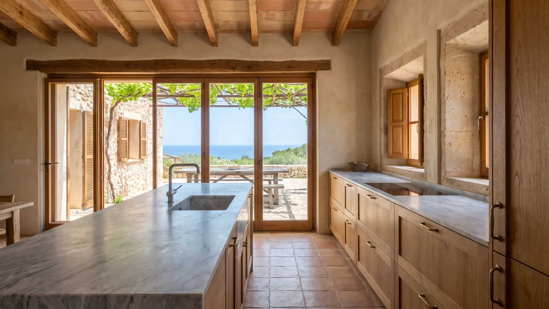 Villa-Küche mit einer grau geäderten Marmor-Kücheninsel und Eichenmöbeln, verglaste Türen zu einem Terrassentisch und dem Meer