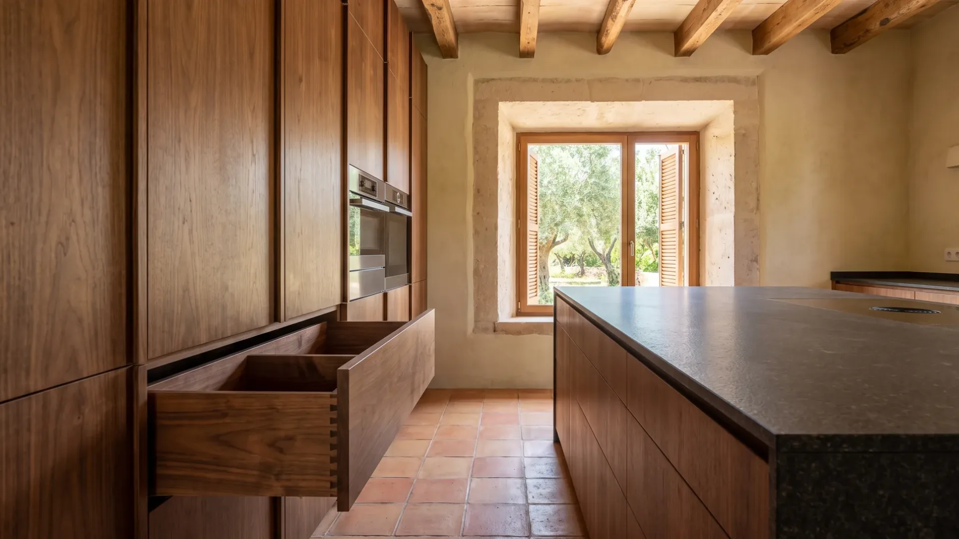 Walnut floor-to-ceiling cabinetry with a deep dovetailed drawer open, a dark granite island and a window onto olive trees