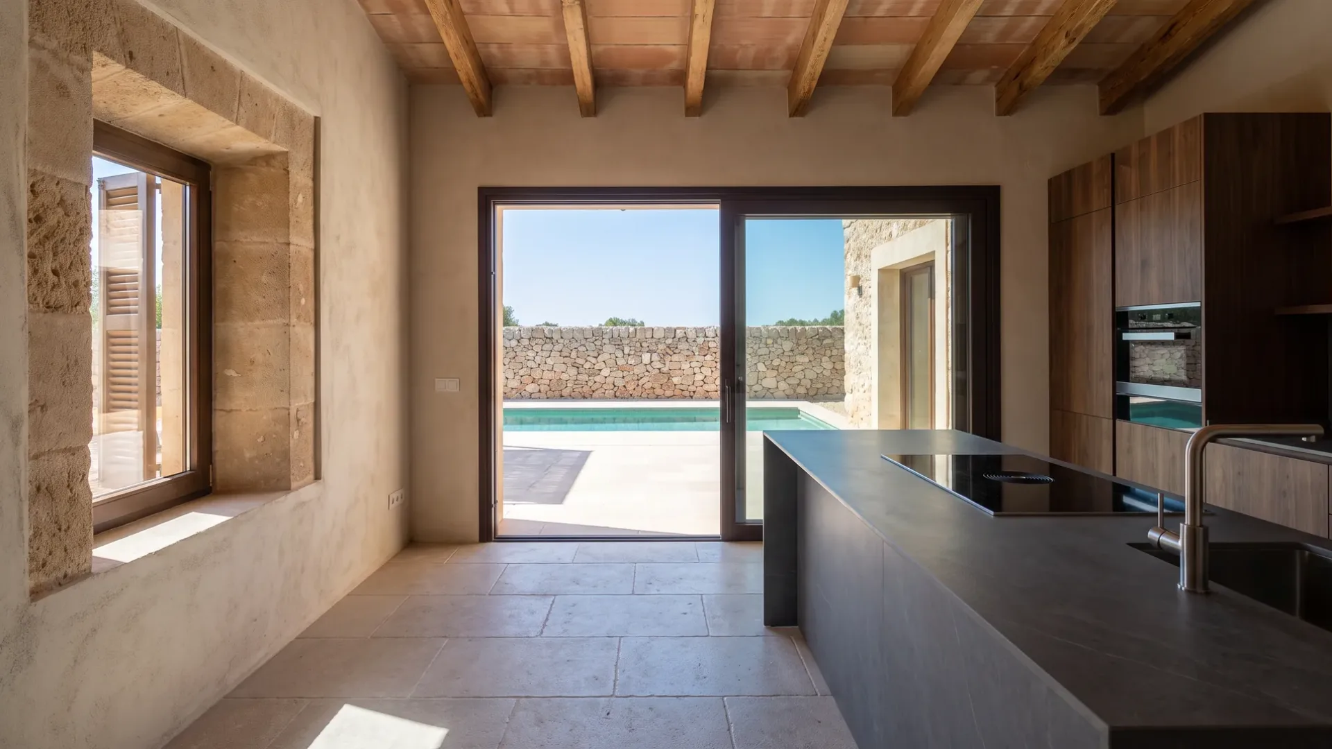 Kalksteinboden, der durch offene Glasschiebetüren zu einer Terrasse mit Pool verläuft, dunkle Keramikinsel und Hochschränke aus Nussbaum