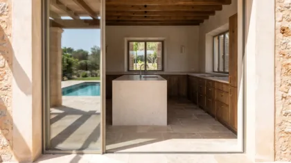 Villa kitchen with a limestone island and walnut units, seen through open glass doors from a pool terrace