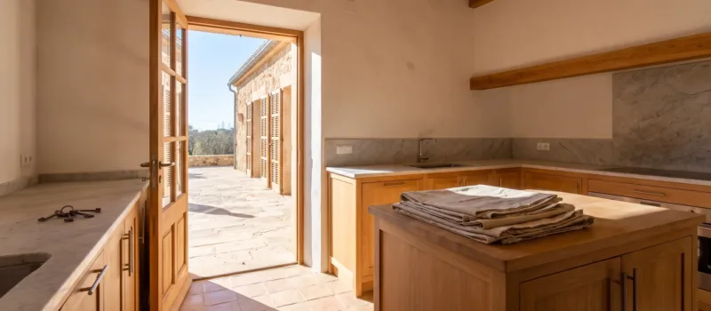 Cocina de roble con encimera de mármol, llaves de la casa y lonas protectoras dobladas sobre la isla, puerta abierta a una terraza soleada