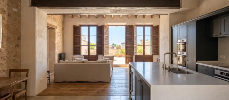 Offene Küche mit dunklen Schränken und einer weißen Steininsel gegenüber einem Wohnzimmer mit Sofa und Türen zu einer Terrasse