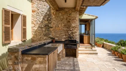 Terrassenküche aus Bruchstein mit dunkler Arbeitsplatte, einer Spüle und integriertem Grill neben einer Steinmauer, dahinter das Meer