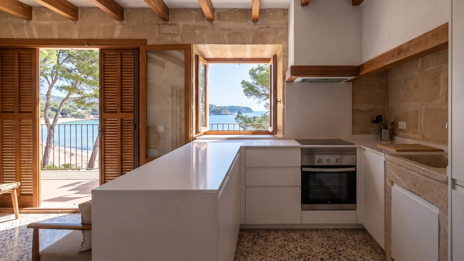 Weiße Küche mit Stein-Arbeitsplatte und Terrazzoboden, offene Fensterläden zu einer Terrasse und einer von Pinien gerahmten Bucht