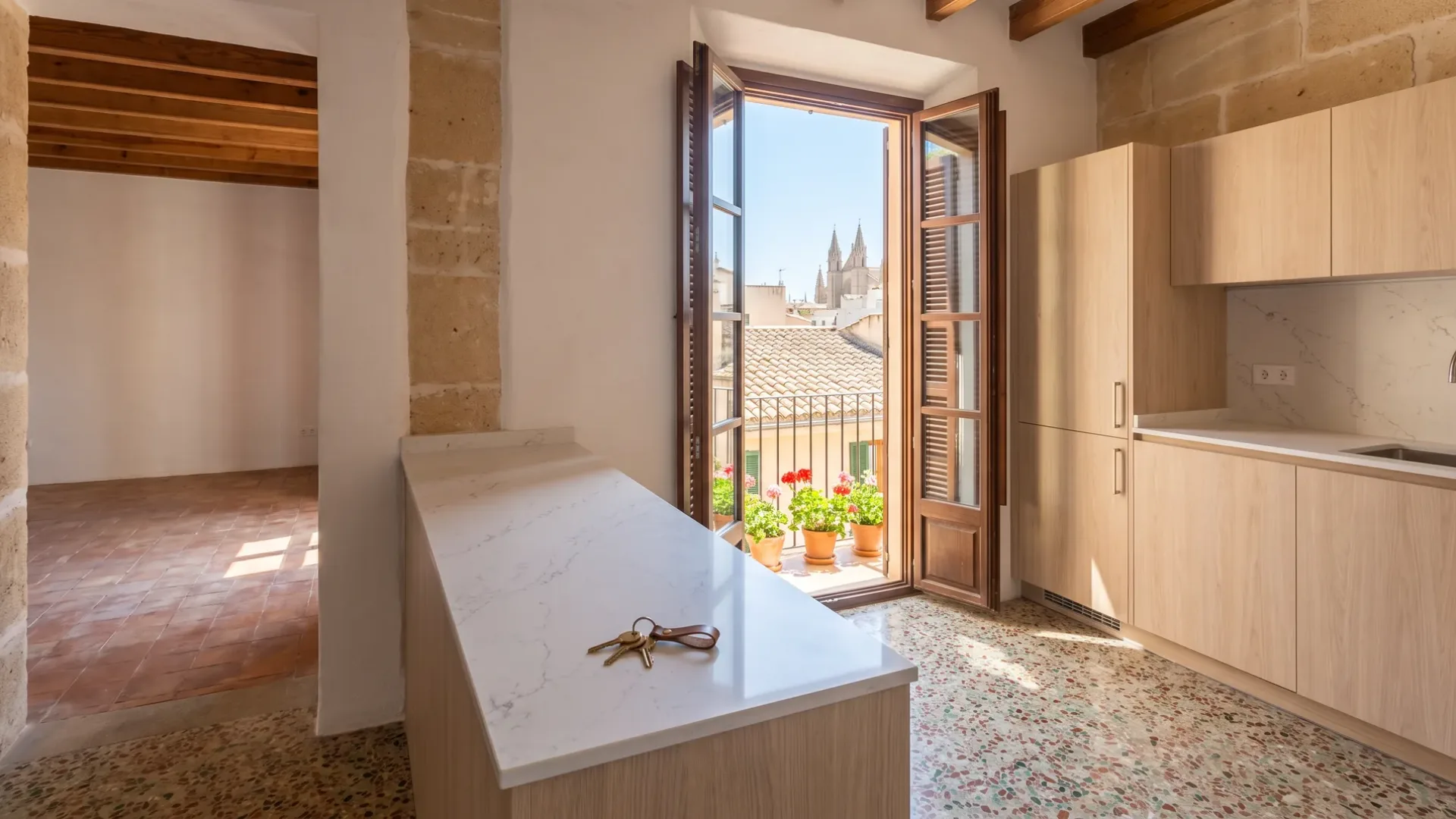 Wohnungsküche aus hellem Eichenholz mit Hausschlüsseln auf weißer Quarzstein-Arbeitsplatte, Terrazzoboden und Balkon über alten Dächern