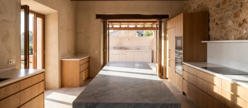 Cocina de villa con isla de mármol veteado en gris, mobiliario de roble y encimeras blancas, puertas abiertas a una cocina exterior