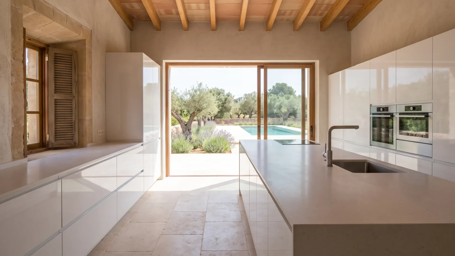 Neue Villa-Küche mit weiß lackierten Schränken, heller Quarzstein-Kücheninsel und Einbaubacköfen, Verglasung zu Lavendel, Oliven und Pool